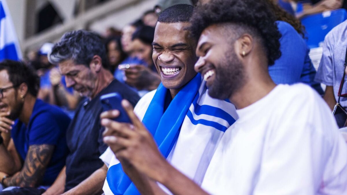 Two young men sit in the stands at a sports event and smile while looking at a phone