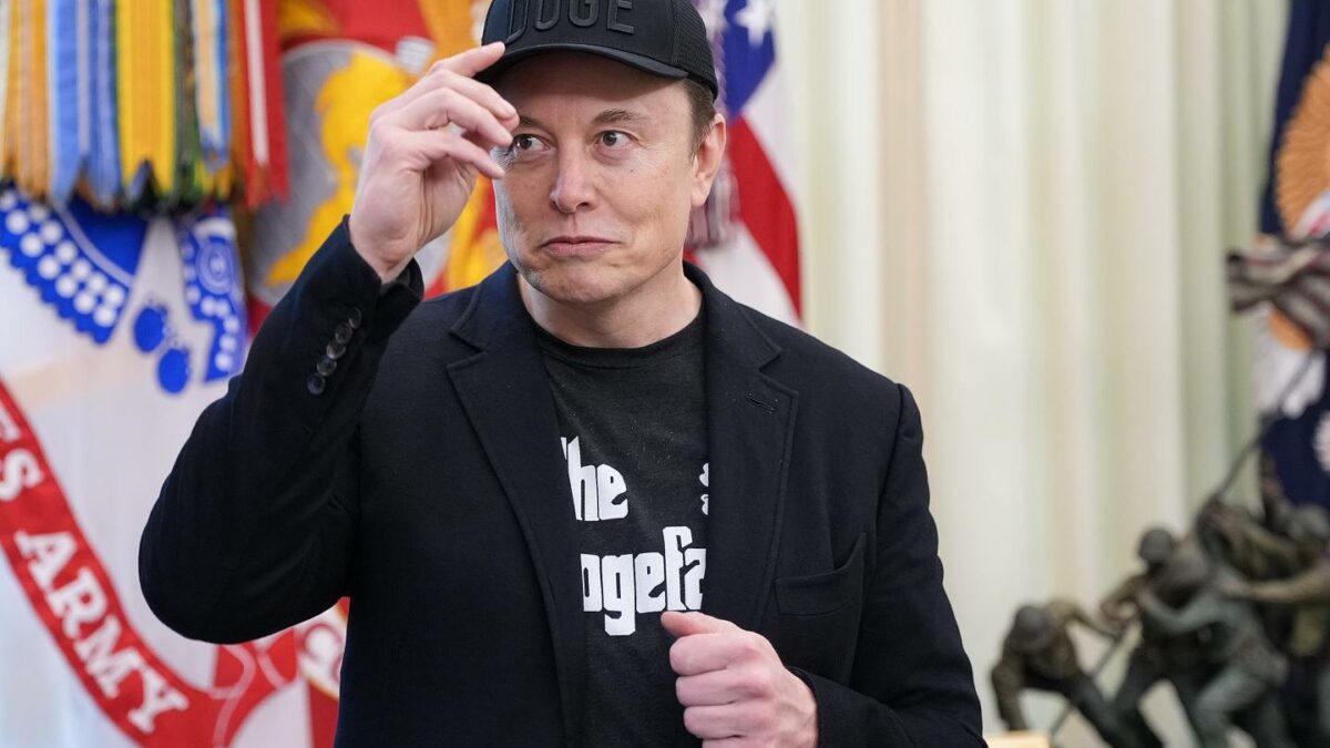 Elon Musk, standing in the oval office, touches an all-black DOGE hat on his head.