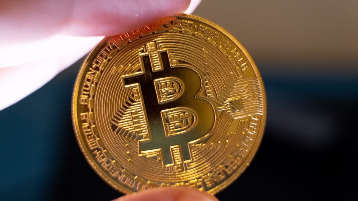 A physical Bitcoin coin held between an index finger and a thumb.