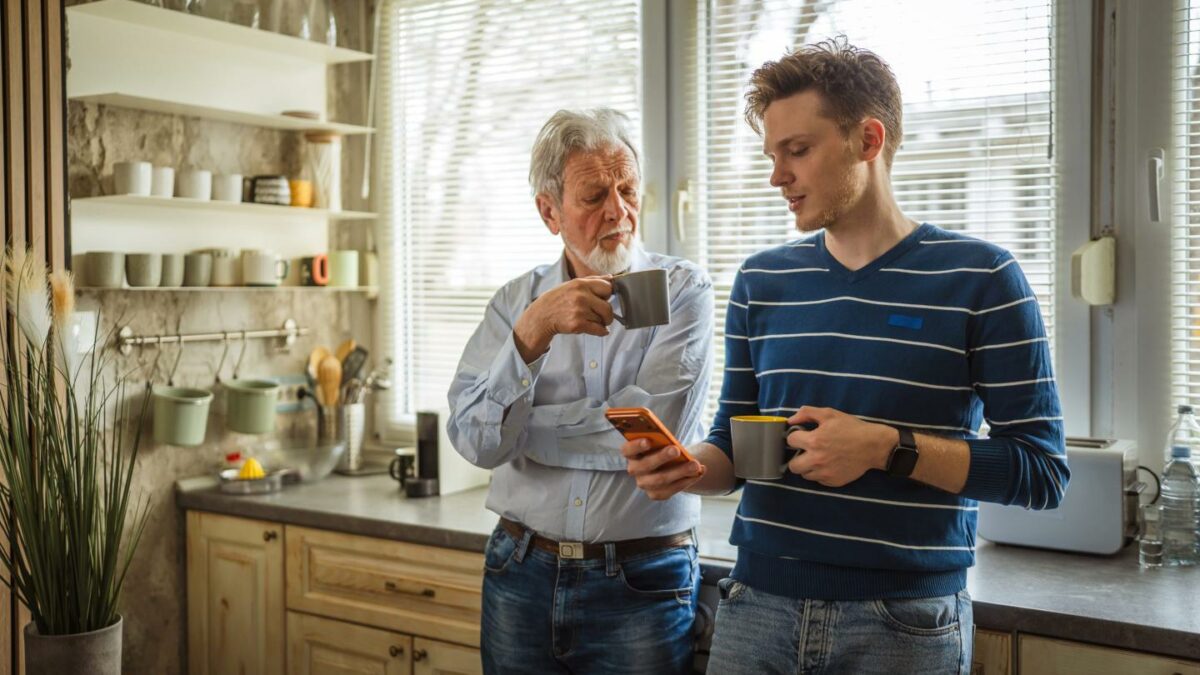 A young man holding a cup of coffee in a kitchen shows his phone to an older man, who is also holding a mug.