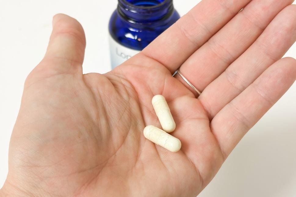 Closeup of an open palm holding two off-white capsules of Healthletic Tri-Active Cellular Longevity Complex. You can see the open blue bottle on the countertop below.
